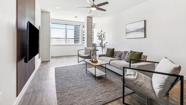 A modern living room with a flat-screen TV, a ceiling fan, a large window, and a minimalist design. - Modern Downtown High-Rise Apartment (Baton Rouge)
