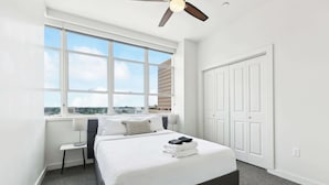 A bedroom with a large window, a bed, a bedside table, a ceiling fan, and a wardrobe.