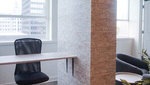 A modern office space with a white brick column, a desk with a black office chair, and a view of the city through large windows.