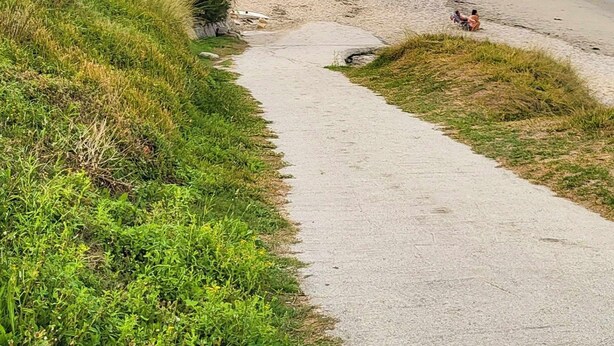 Camino costero que lleva a una playa con aguas azules cristalinas y un paisaje montañoso.