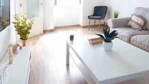 Una sala de estar moderna con un sofá blanco, una mesa de centro y un televisor sobre un mueble blanco. Se ve una puerta que da a otra habitación y una ventana con persianas.