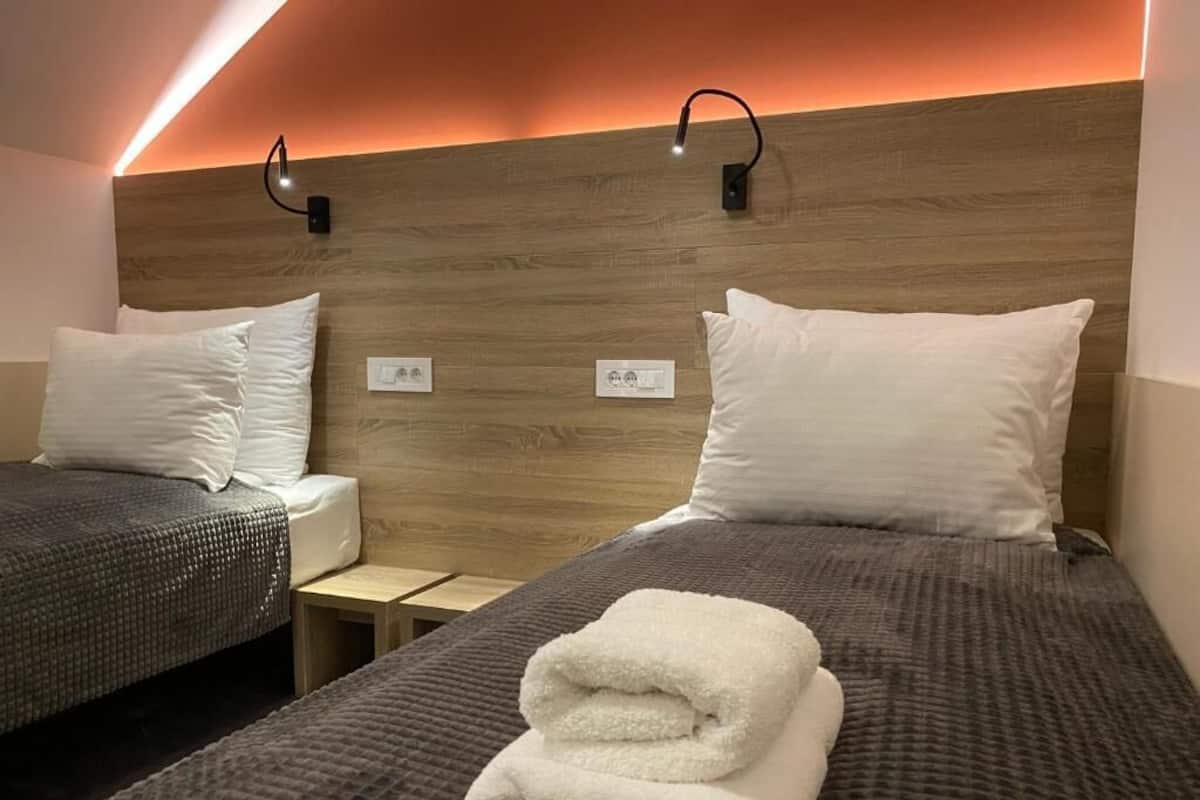A hotel room with two beds, each with a pillow and a folded towel. There are two bedside tables, one on each bed. The room has a wooden headboard with a light strip running along the top.