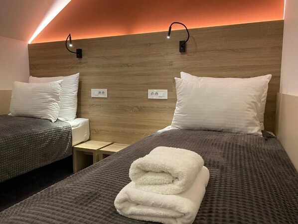 A hotel room with two beds, each with a pillow and a folded towel. There are two bedside tables, one on each bed. The room has a wooden headboard with a light strip running along the top. - Konak Stella (Belgrade)