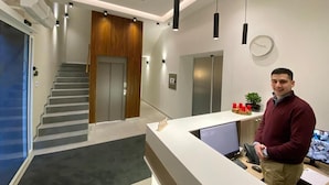 A modern reception area with a desk, computer, and a person standing. There is a staircase and a clock on the wall. - Konak Stella (Belgrade)