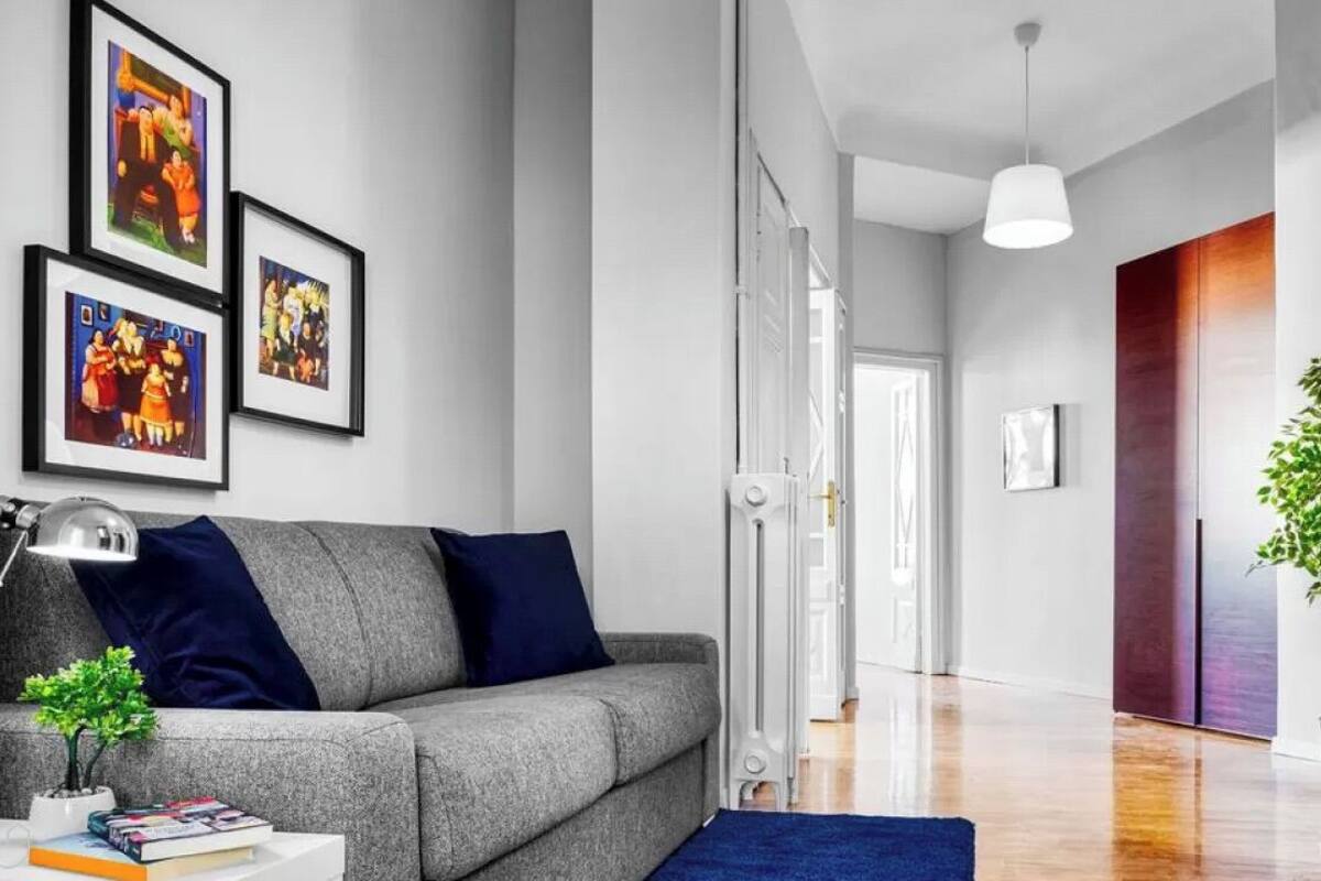 A modern living room with a grey sofa, blue cushions, a small table with books and a plant, and framed artwork on the wall.