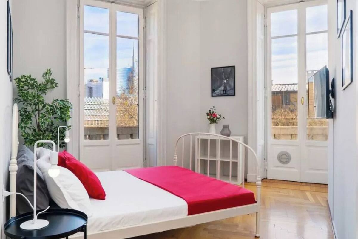 A bedroom with a bed, a bedside table, a potted plant, and a window with a city view.