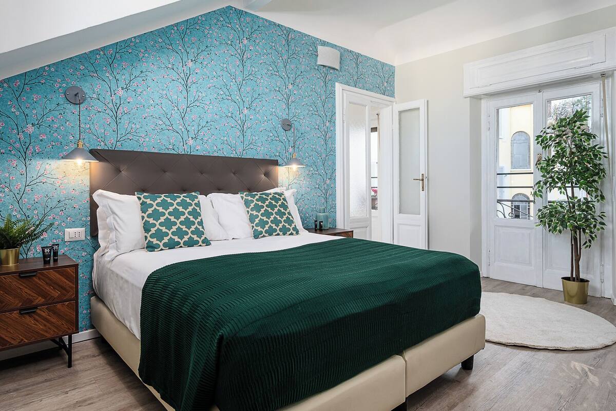 A bedroom with a bed, nightstands, a dresser, a potted plant, and a view of the outside.