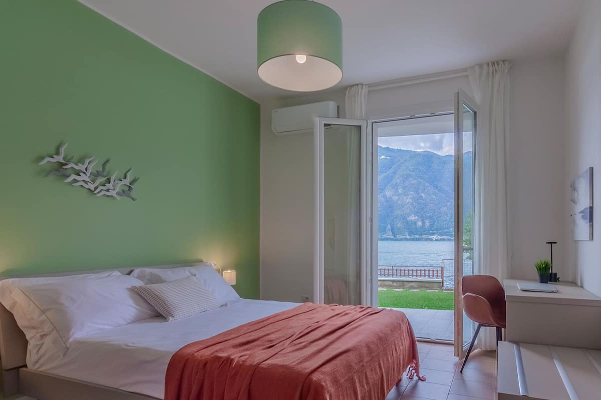 A bedroom with a bed, a desk, and a view of a lake and mountains.