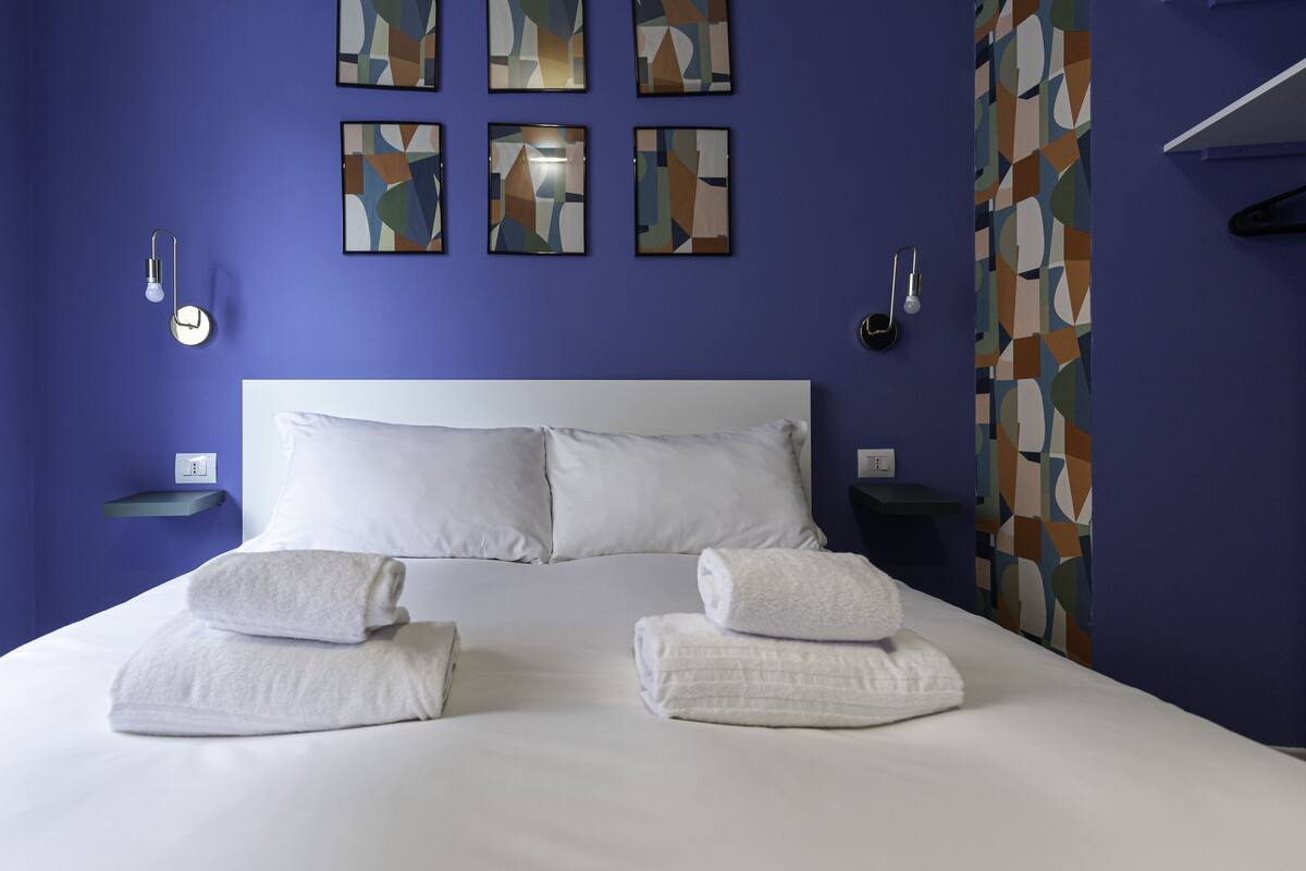 A hotel room with a large bed, white linens, two bedside tables, a wall-mounted light, and abstract artwork.