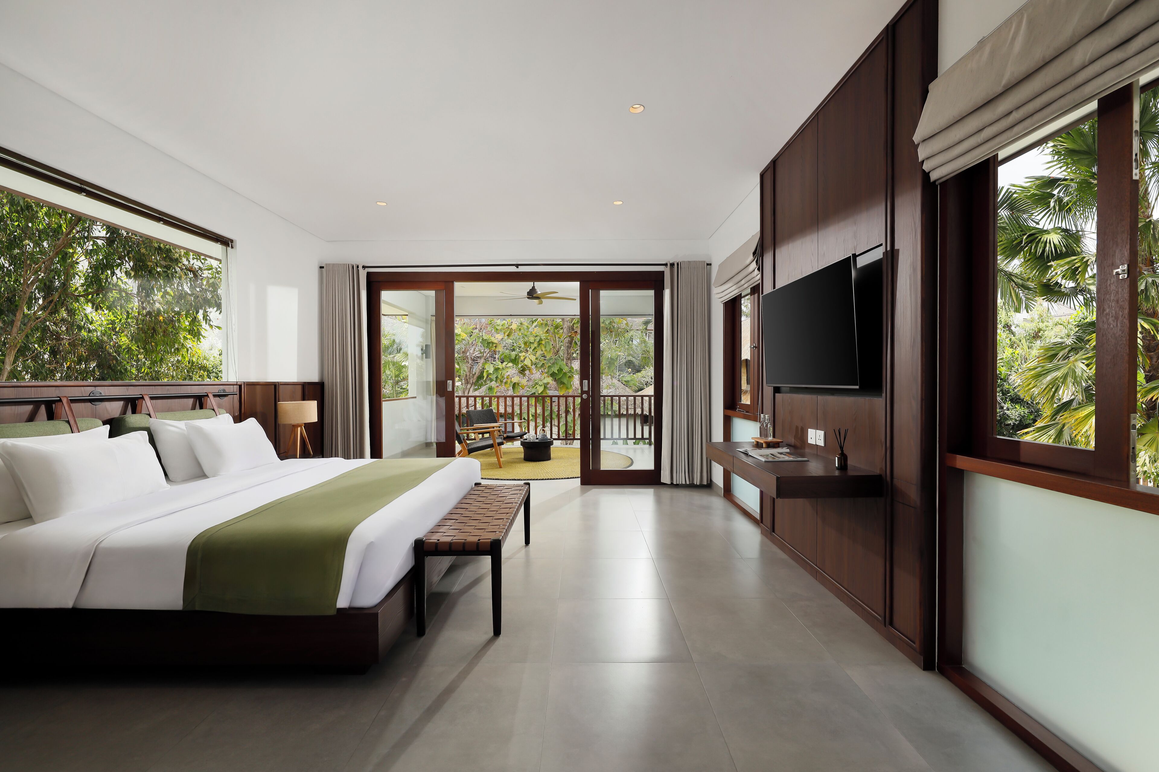A modern hotel room with a large bed, a flat-screen TV, and a balcony with a view of lush greenery.