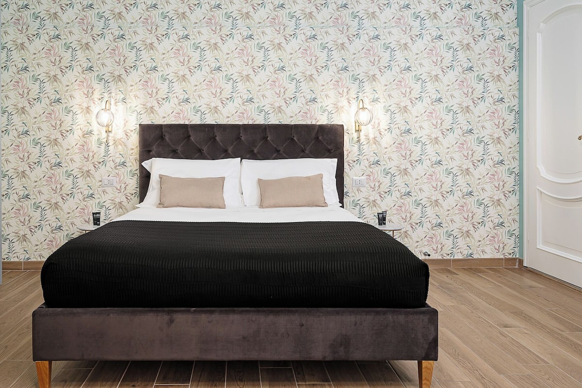 A modern bedroom with a tufted headboard, a black bedspread, and a floral wallpaper pattern.