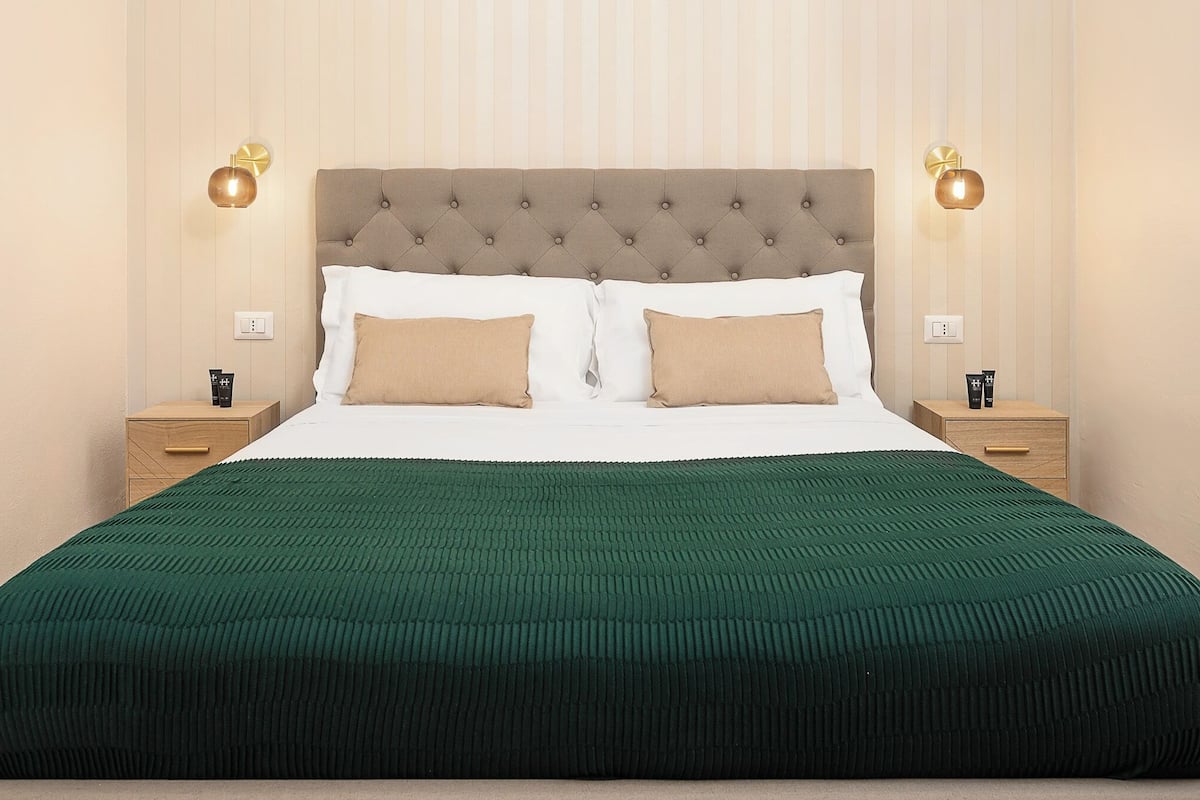 A neatly made bed with a green bedspread, two pillows, and a tufted headboard. There are two bedside tables with lamps and a nightstand with a lamp.