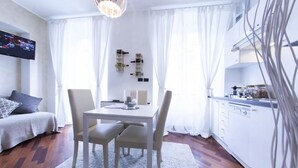 Un soggiorno moderno con zona pranzo, divano, TV e cucina con mobili e piano di lavoro bianchi.