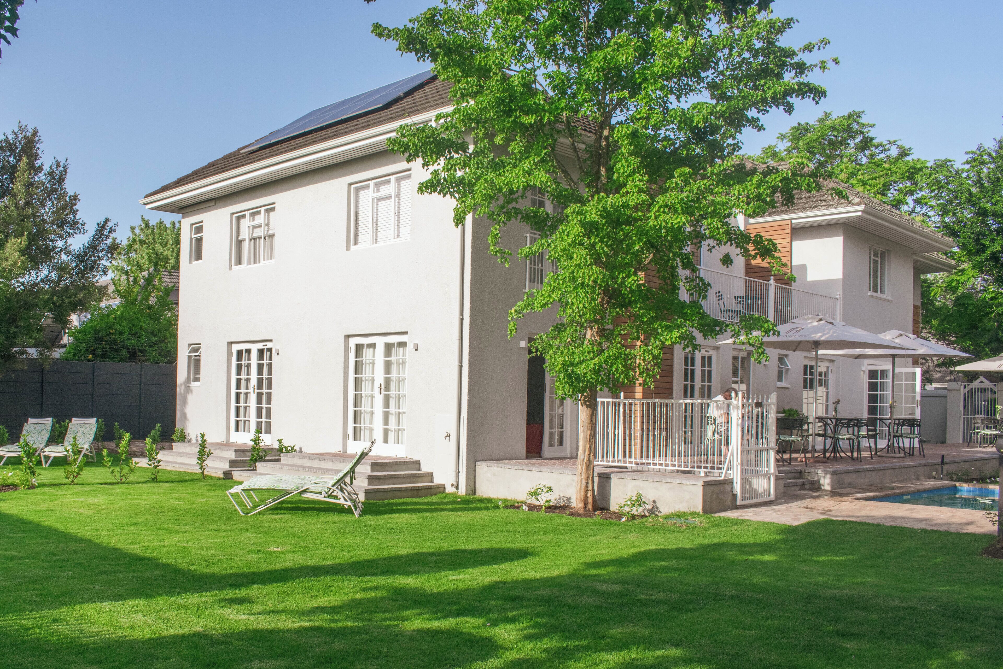 A two-story house with a swimming pool, a patio with outdoor furniture, and a tree in the yard.