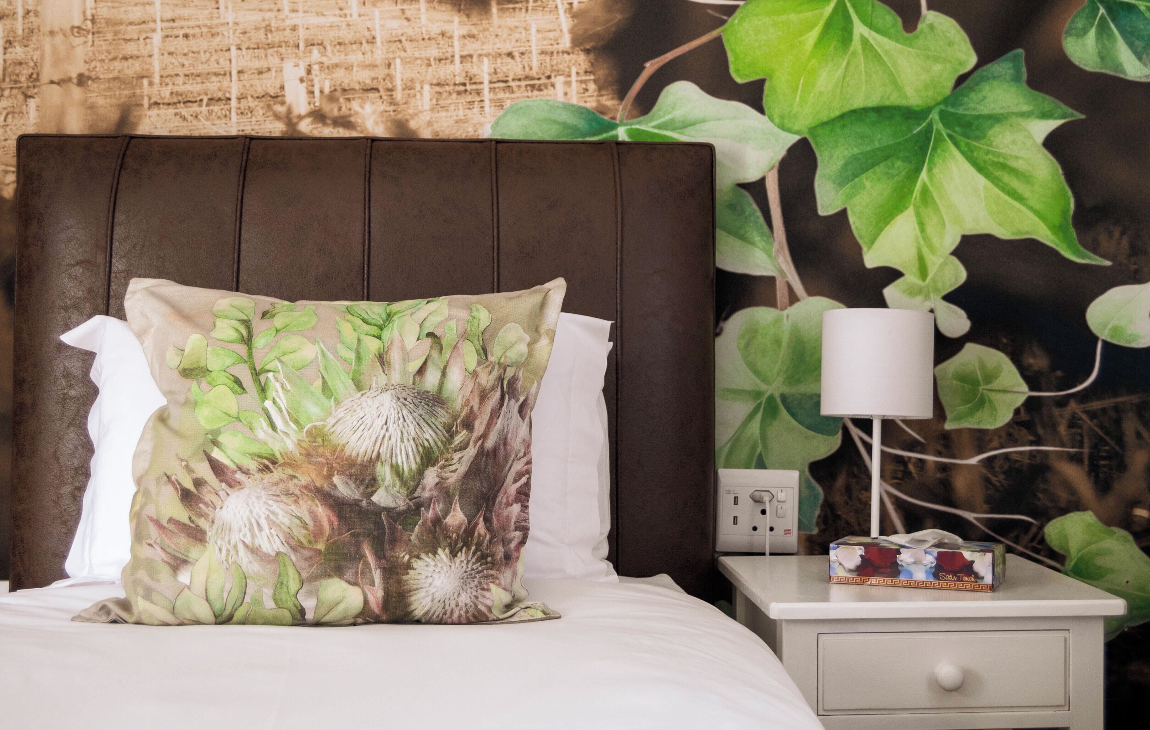 Una cama con un cojín floral, una mesita de noche con una lámpara y una pared con un mural de hojas.