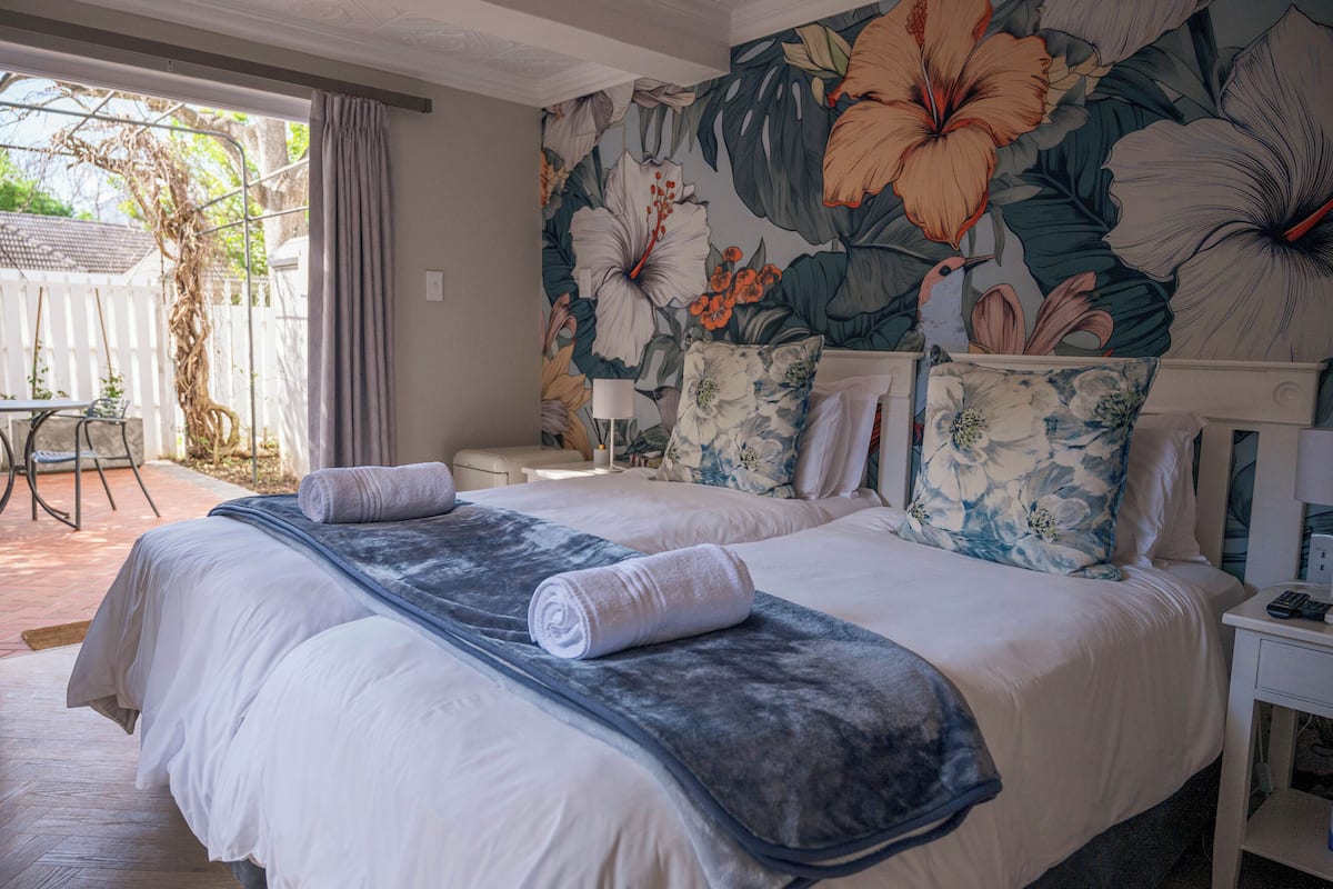 Una cama doble con sábanas blancas y una manta azul, una mesita de noche con lámpara, y un mural floral en la pared.