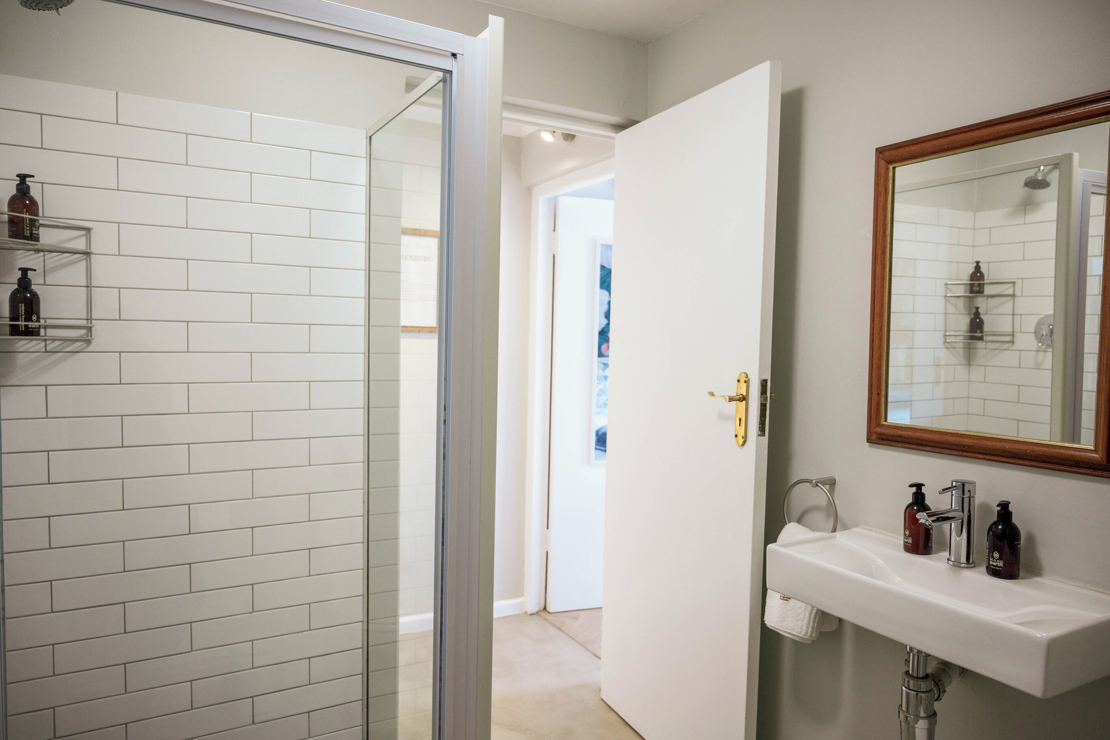 Et badeværelse med hvide flisemure, en bruseglassdør, en hvid vask med et spejl i træramme og to hylder med toiletartikler.