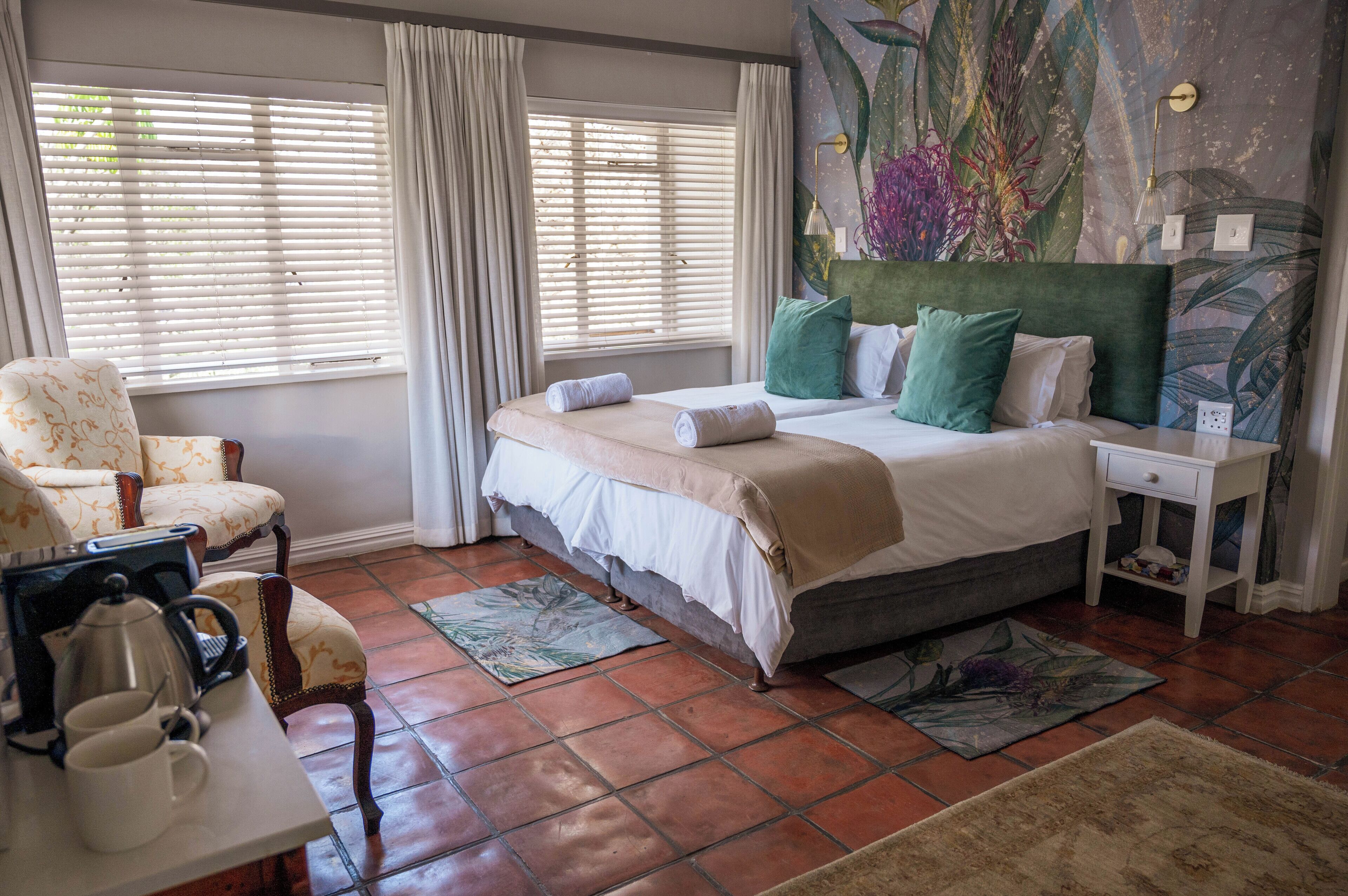 A bedroom with a bed, a chair, a nightstand, a table with a kettle, and a window with curtains.