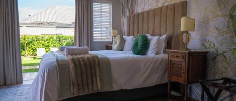 Un dormitorio con una cama grande, una mesita de noche, una lámpara y vistas al exterior con vegetación.