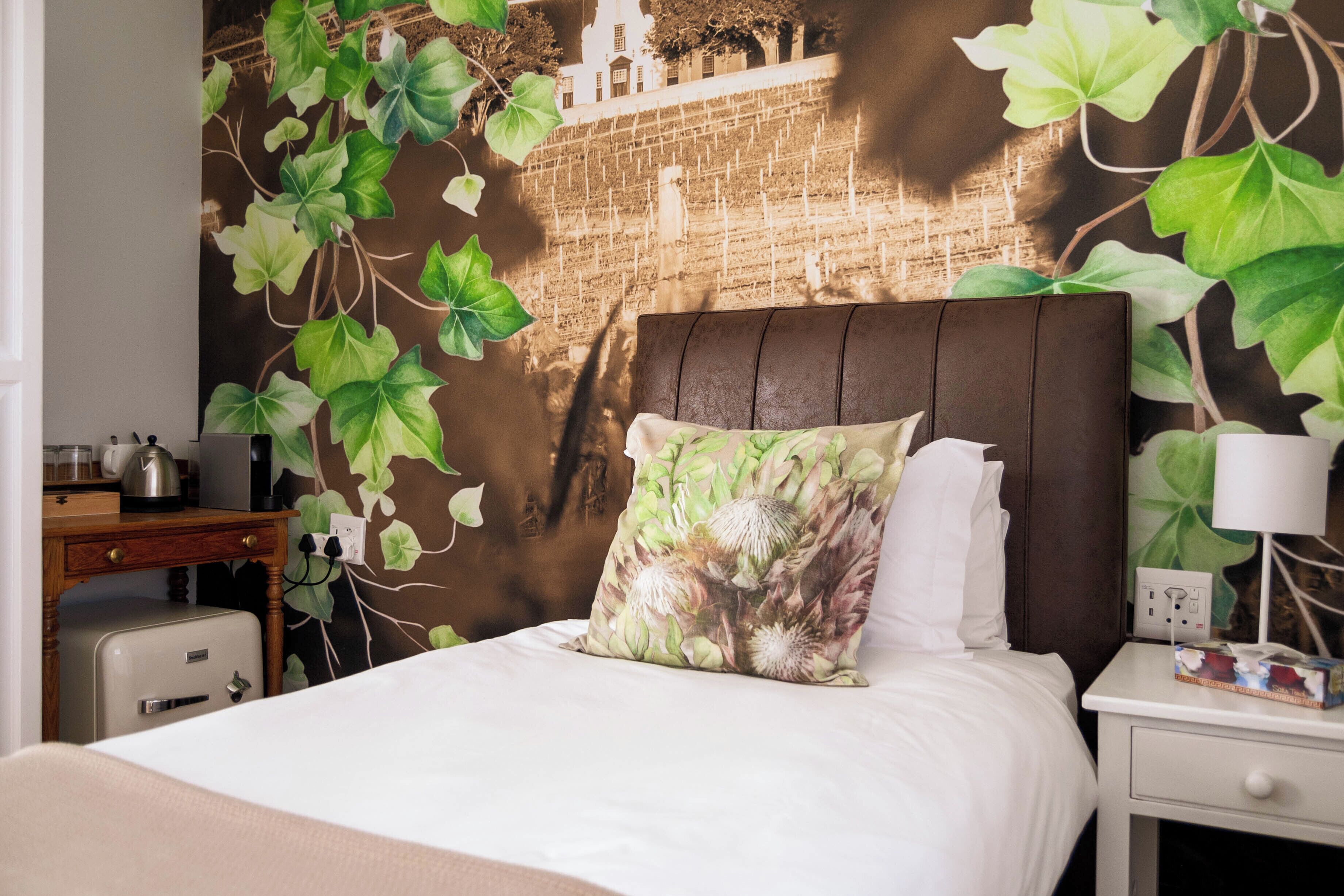 Uma cama com travesseiro floral, uma mesa de cabeceira com abajur e uma parede com mural de hera.