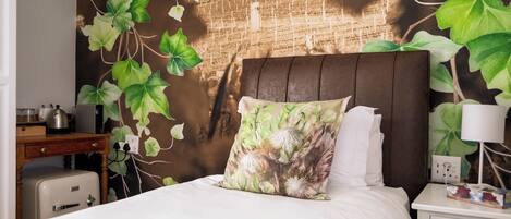 Uma cama com travesseiro floral, uma mesa de cabeceira com abajur e uma parede com mural de hera.
