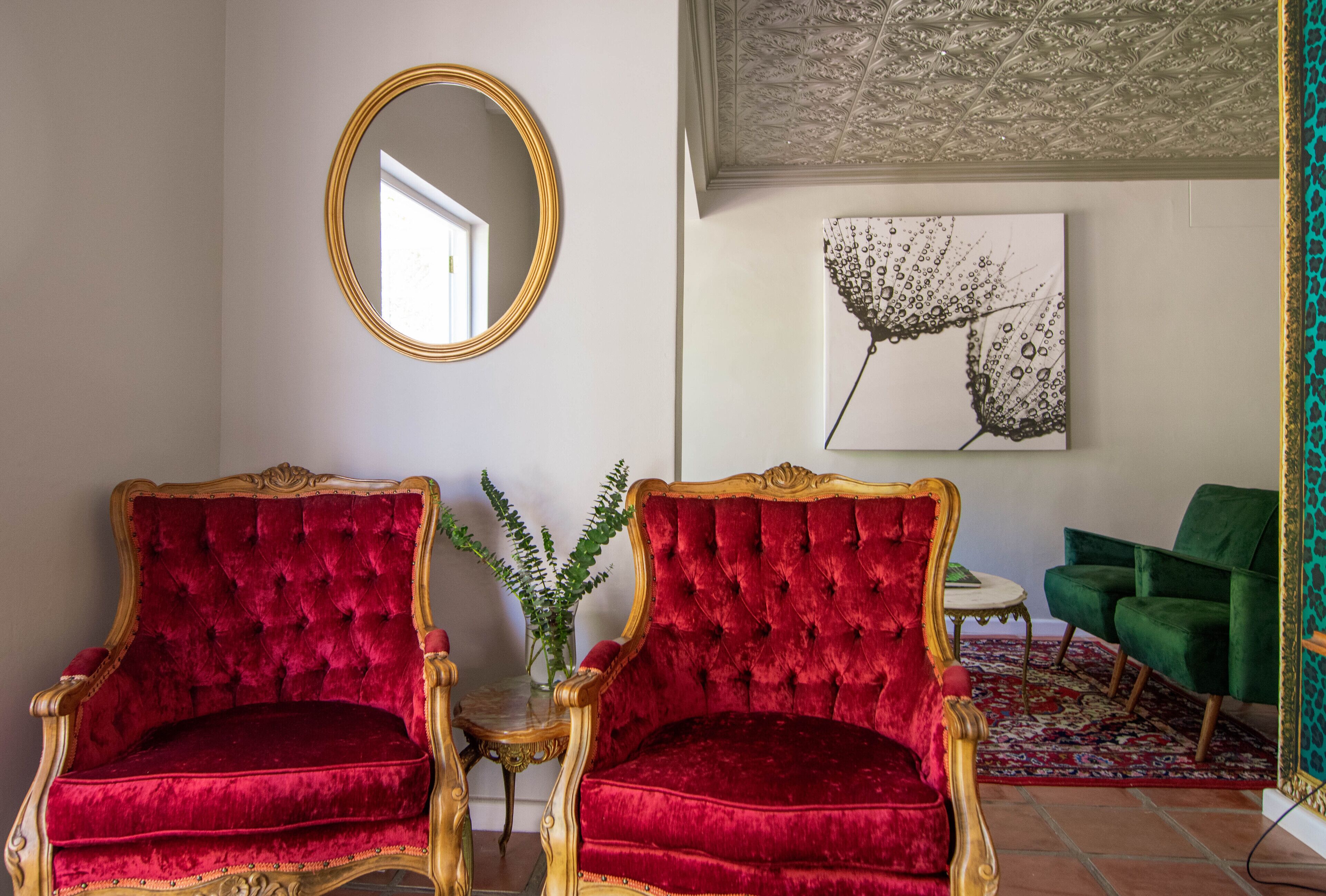 A room with two red velvet armchairs, a green armchair, a round mirror, and a framed artwork on the wall.