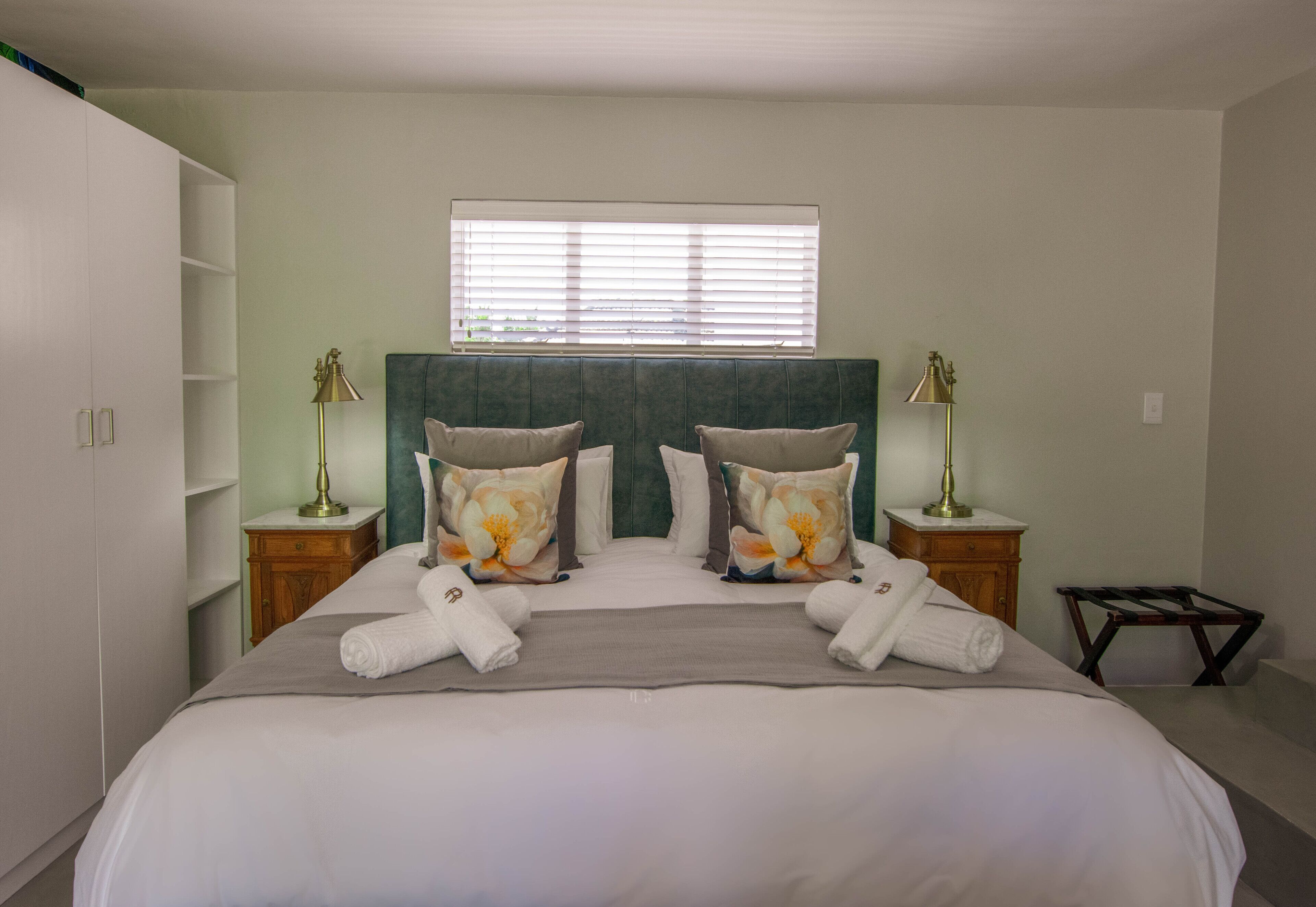 A neatly made bed with two pillows, a bedside table with a lamp, and a window with blinds.