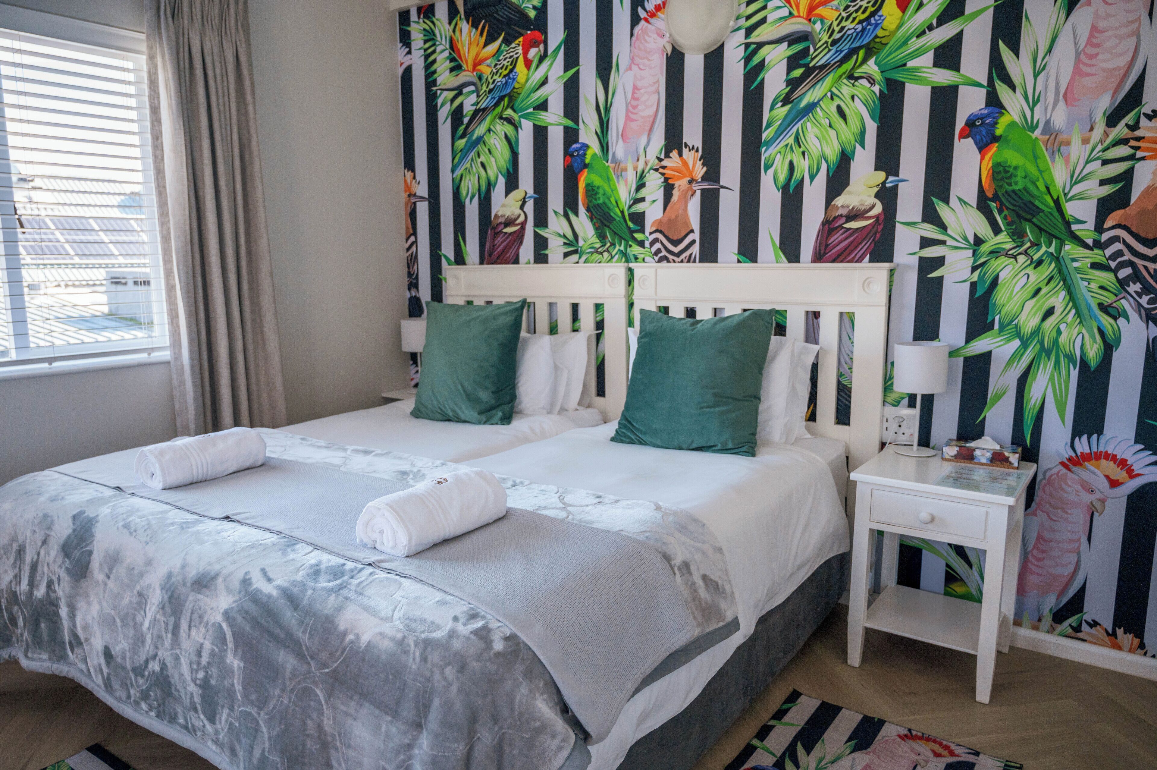 A double bed with green and white bedding, a nightstand with a lamp, and a wall with tropical bird wallpaper.