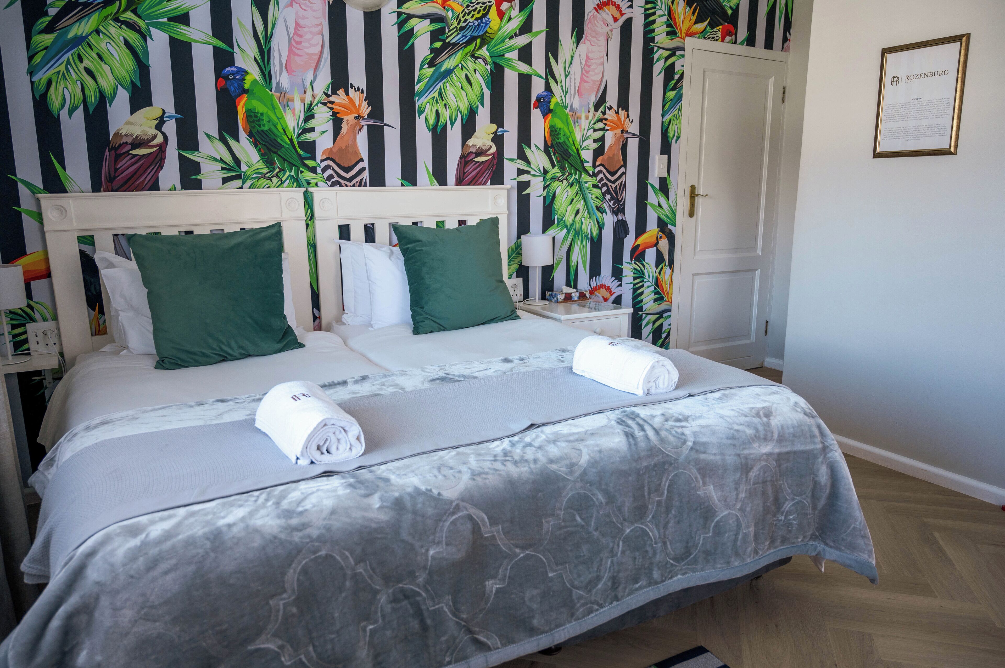 A double bed with green pillows, a grey patterned bedspread, and two rolled towels.