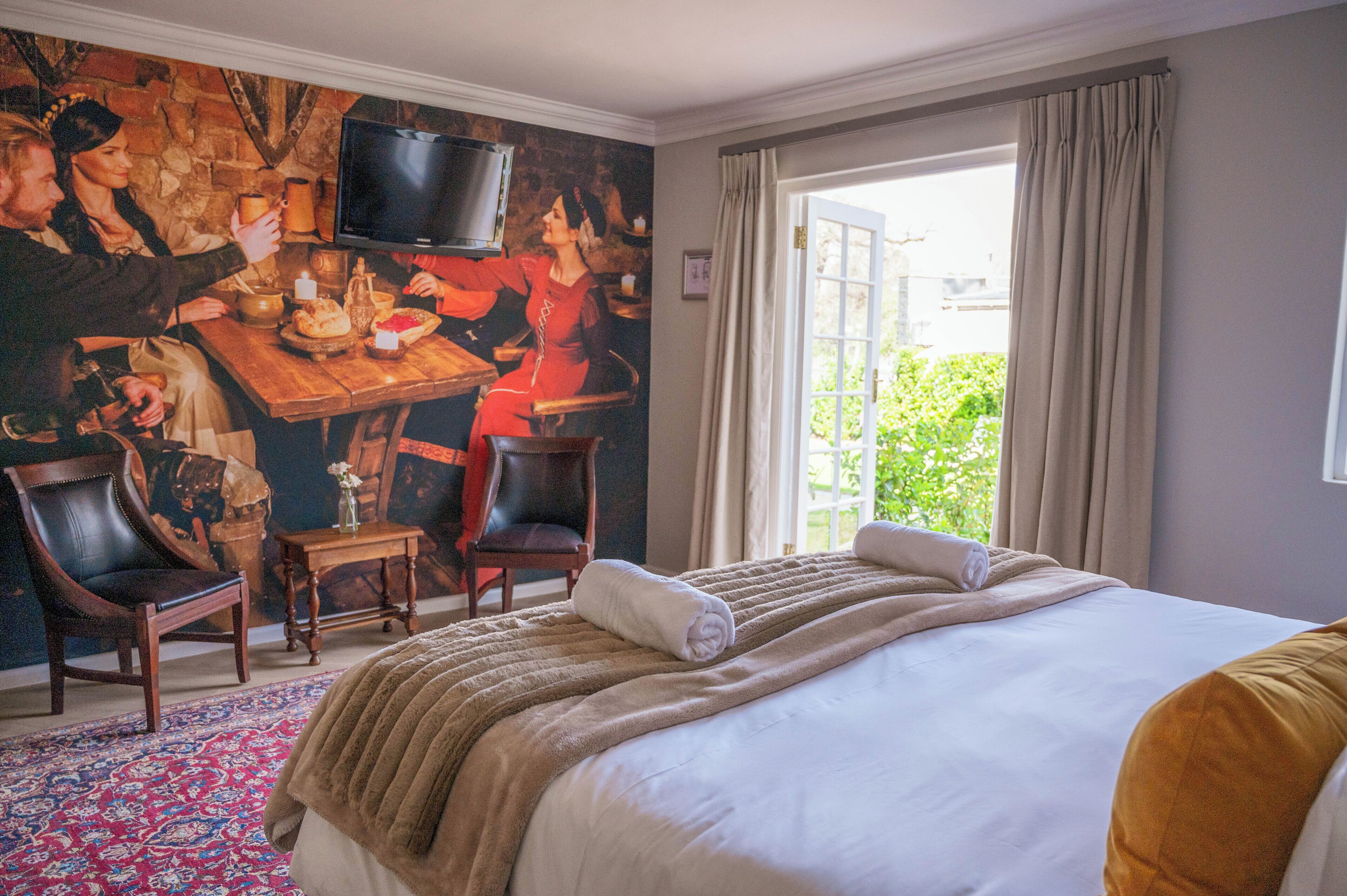 Habitación de hotel con una cama grande, televisión, mesa de comedor con sillas y vistas al exterior con vegetación.