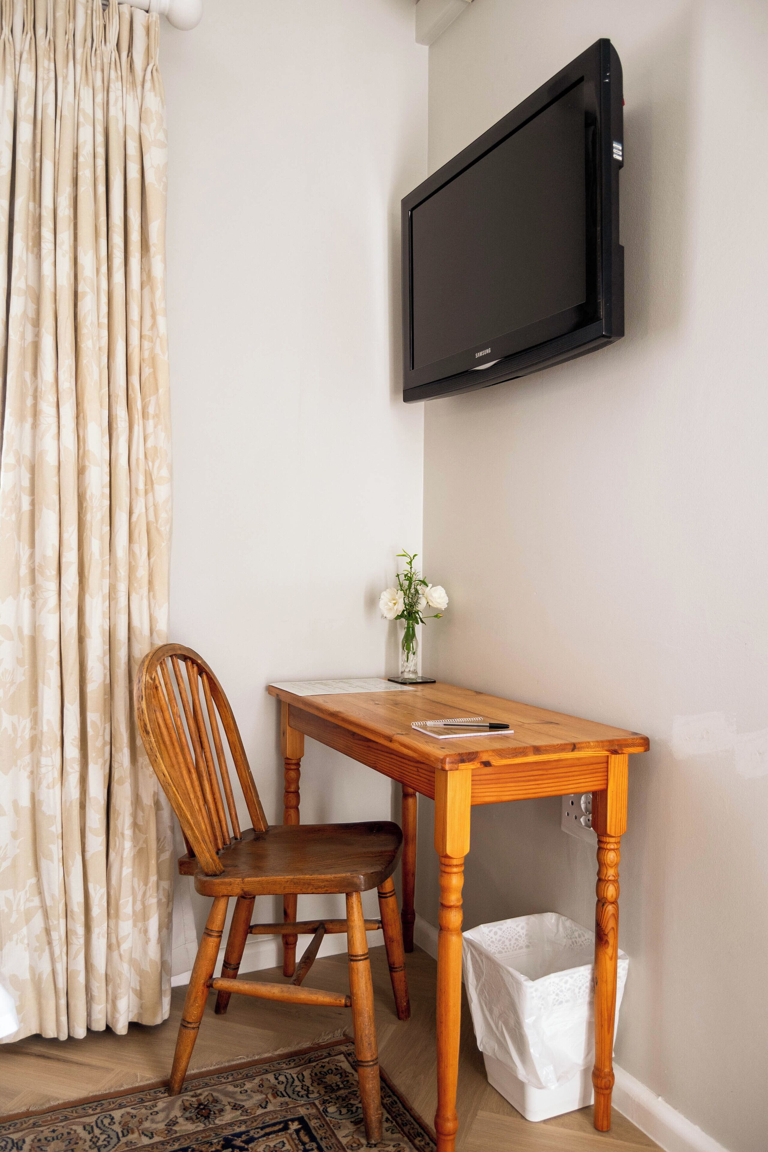 A room with a wooden table, a chair, a TV mounted on the wall, a white trash bin, and a curtain.