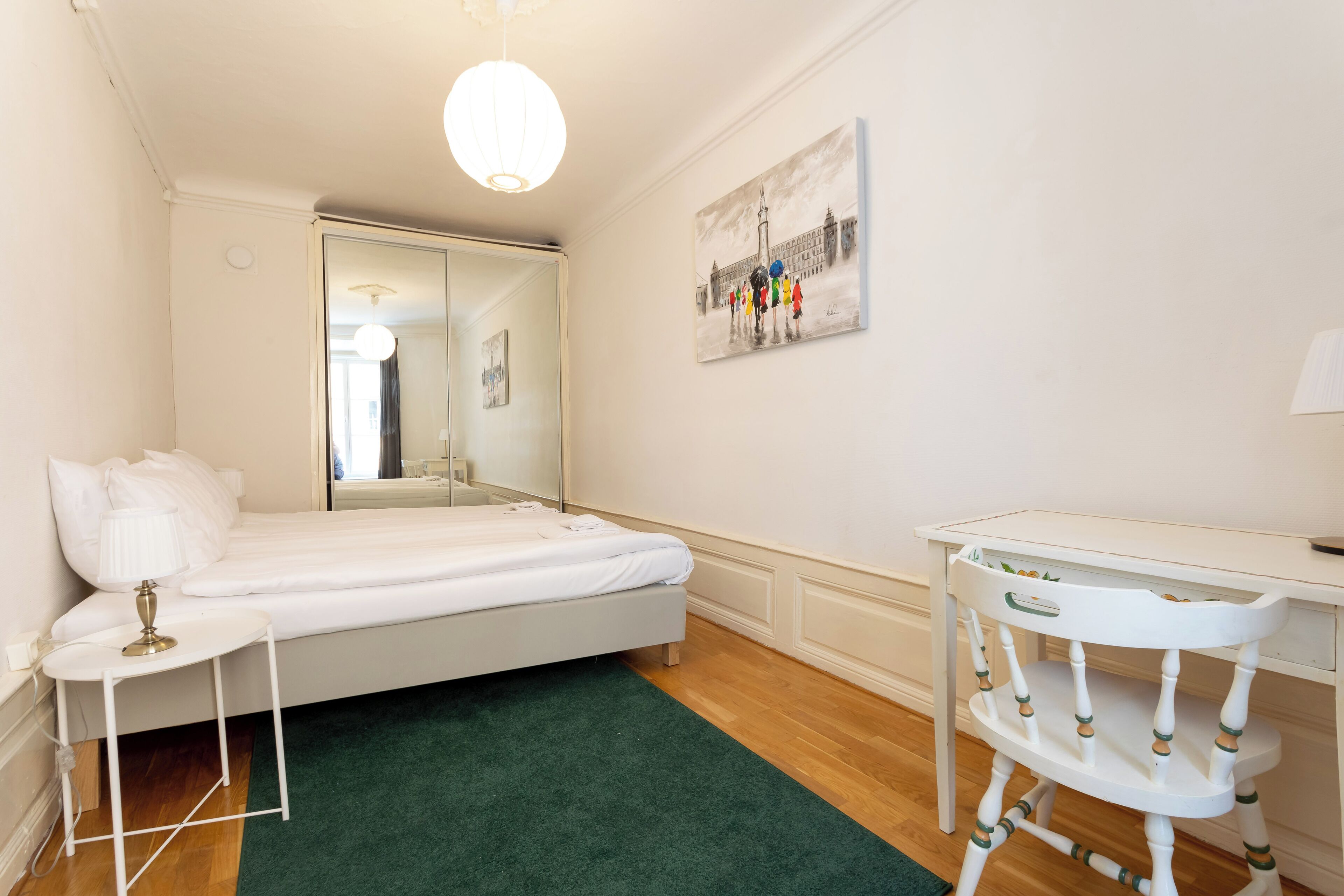 A bedroom with a bed, a desk, a chair, a green rug, and a painting on the wall.