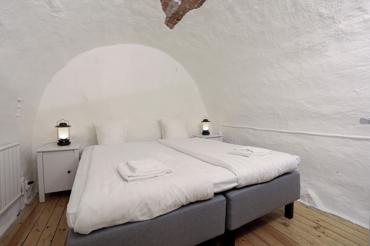 Un dormitorio con cama blanca, cabecera gris y dos mesitas de noche con faroles.