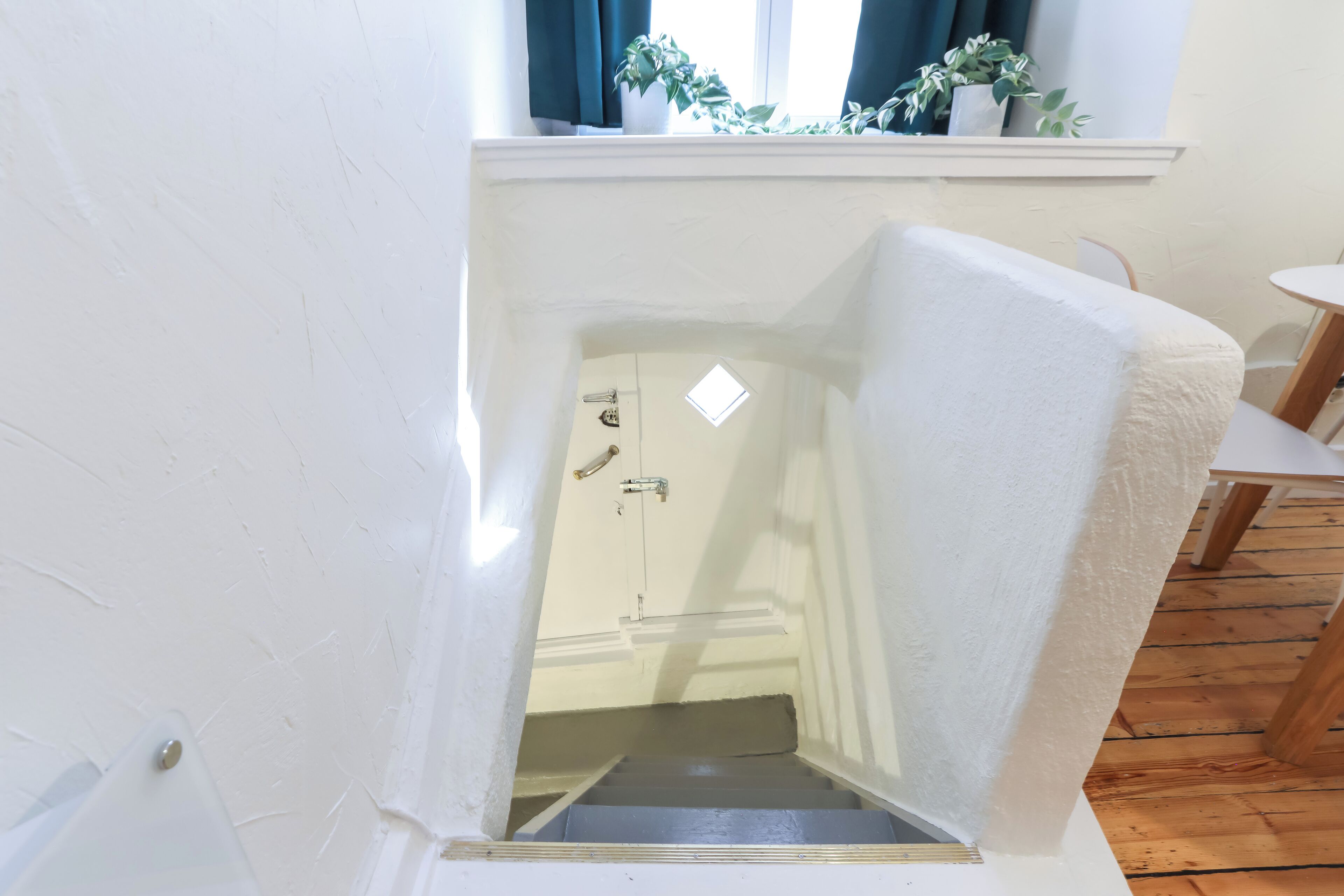 Een kamer met een witte muur, een raam met gordijnen, een kleine plant in een pot en een houten vloer.