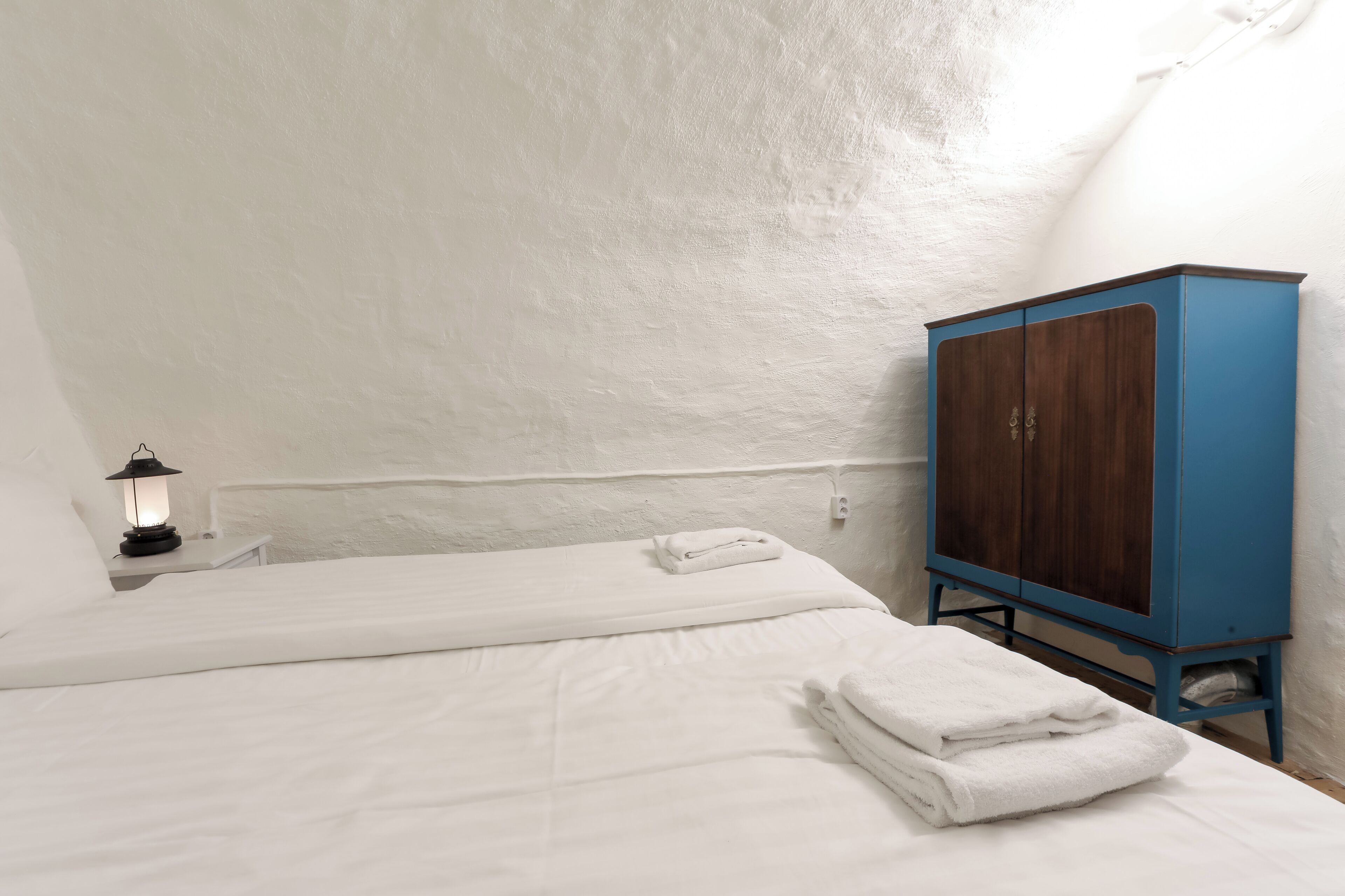 A single bed with white linens and a folded towel, a small bedside table with a lantern, and a blue cabinet against a white wall.