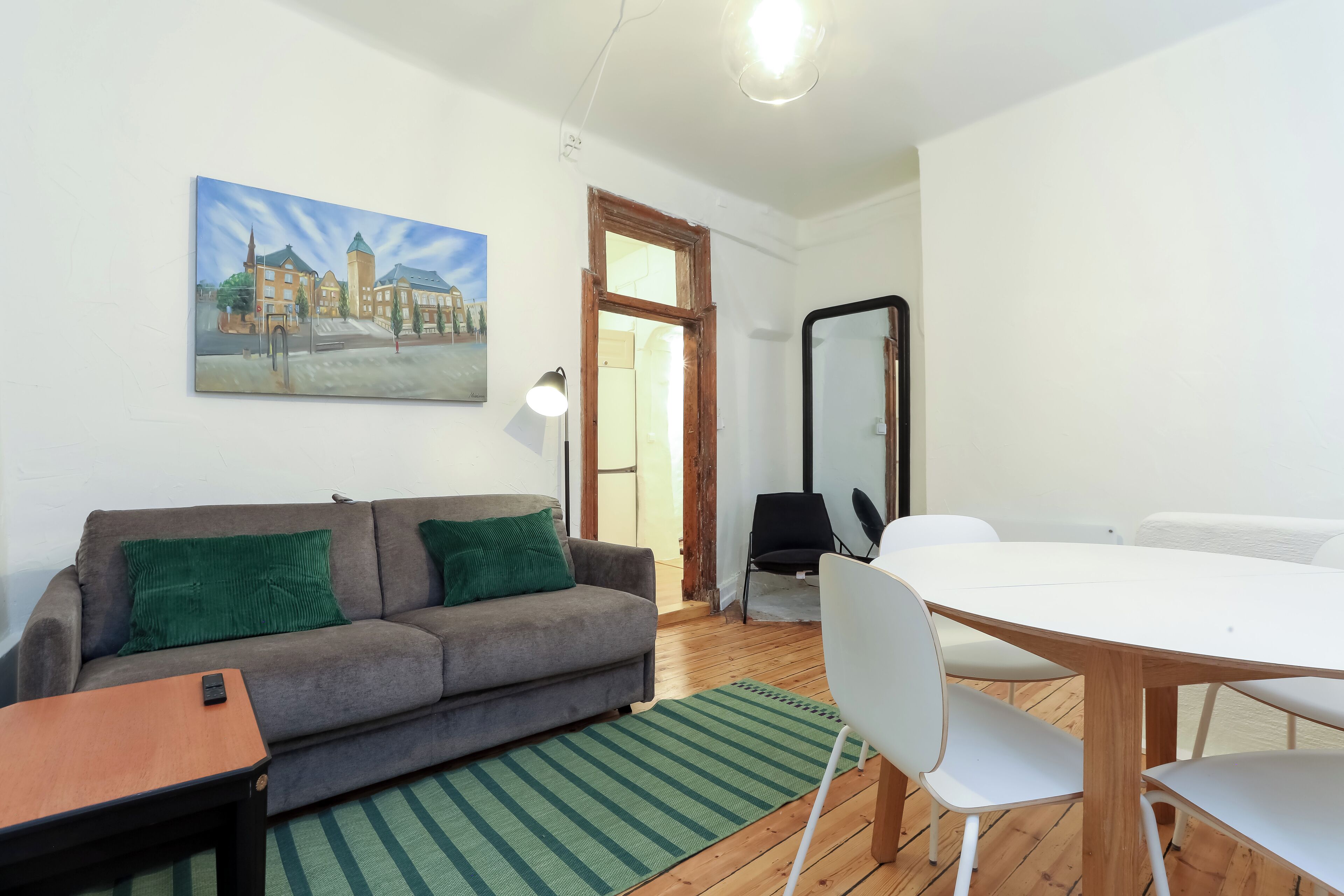 Un salon moderne avec un canapé gris, des coussins verts, une petite table en bois et une table à manger blanche avec des chaises. Un tableau représentant un bâtiment est accroché au mur, et un miroir se trouve sur le mur opposé au canapé.