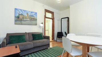 Un salon moderne avec un canapé gris, des coussins verts, une petite table en bois et une table à manger blanche avec des chaises. Un tableau représentant un bâtiment est accroché au mur, et un miroir se trouve sur le mur opposé au canapé.