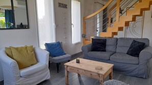 Una sala moderna con un sofá gris, un sillón blanco y una mesa de centro de madera. En el fondo, se ve una escalera con barandilla de madera.