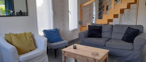 A modern living room with a grey sofa, a white armchair, and a wooden coffee table. A staircase with a wooden railing is visible in the background.