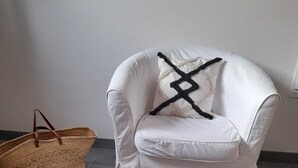 Un sillón blanco con un plaid blanco y negro, una cesta tejida y un reposapiés mullido.