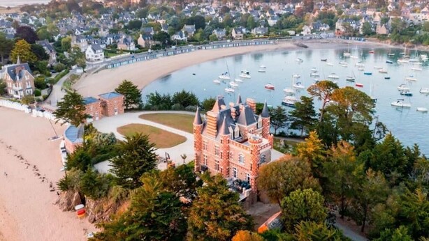 Un pueblo costero con un imponente edificio que recuerda a un castillo, una playa y un puerto deportivo lleno de barcos.