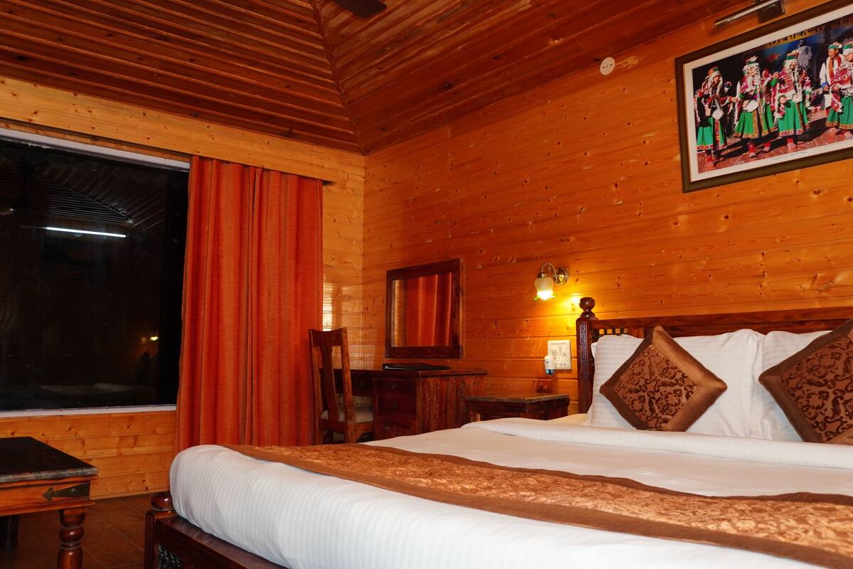 Una habitación de madera con cama, una ventana con cortinas rojas, un cuadro en la pared y una mesita de noche con lámpara.