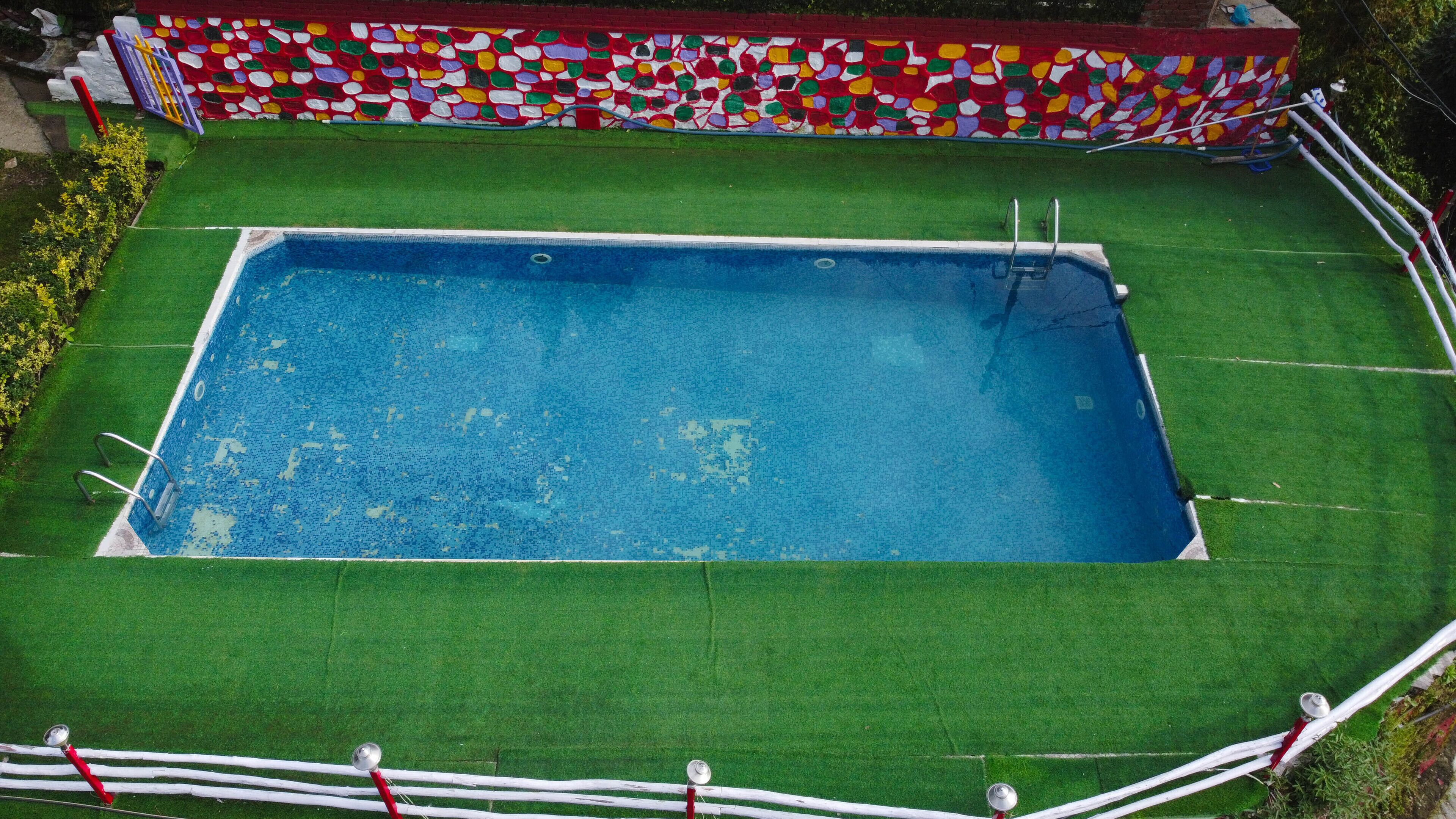 Una piscina rectangular con revestimiento de mosaicos azules, rodeada por un deck de césped artificial verde y una pared de mosaicos de colores.