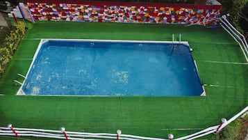 Una piscina rectangular con revestimiento de mosaicos azules, rodeada por un deck de césped artificial verde y una pared de mosaicos de colores.