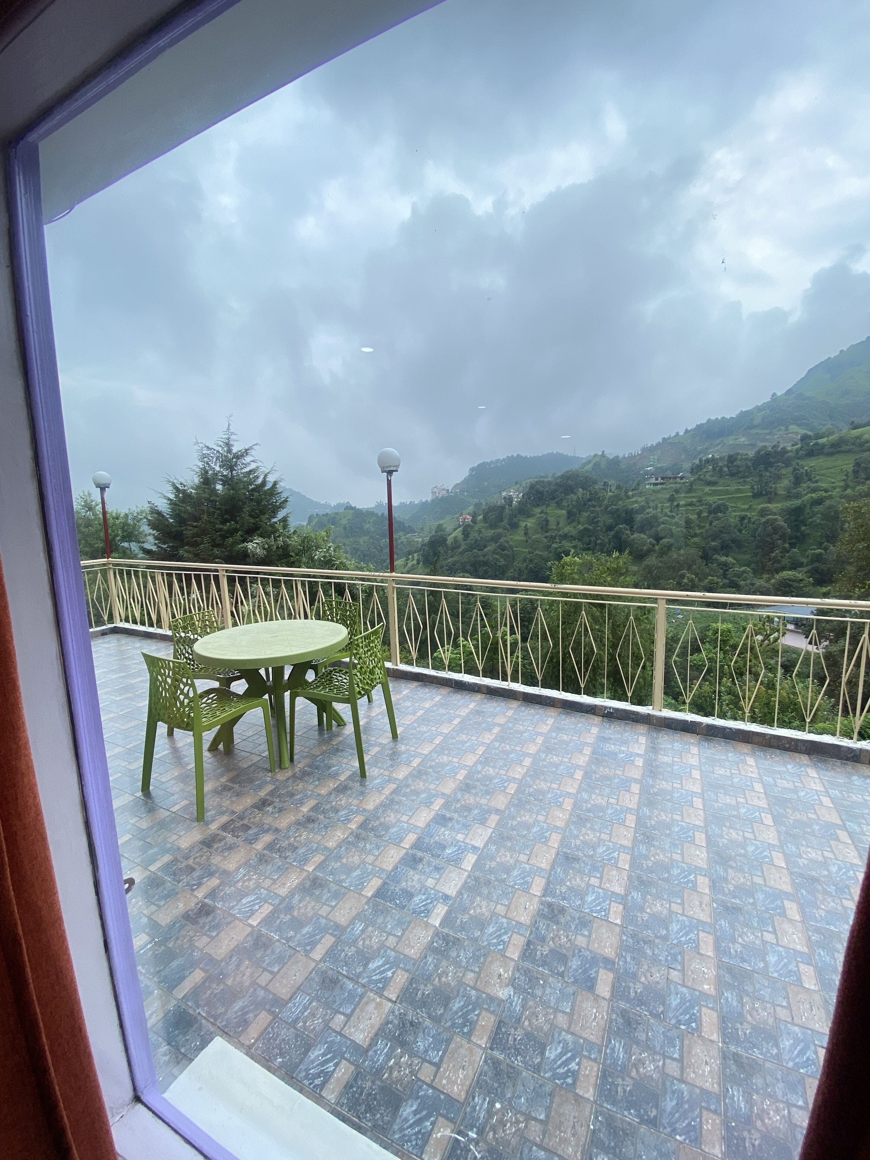 A balcony with a tiled floor, a table with chairs, and a view of a green landscape with hills and trees.