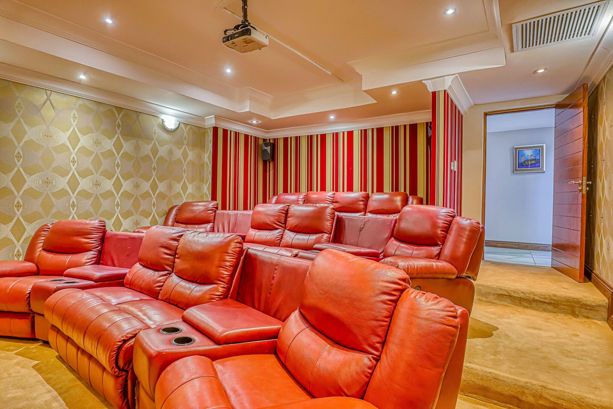 Sala de cinema com poltronas reclináveis de couro vermelho, projetor no teto e papel de parede listrado.