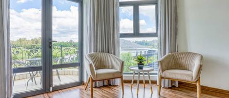 Una habitación con dos sillones color beige, una mesita redonda con una planta en maceta y ventanas amplias con cortinas.