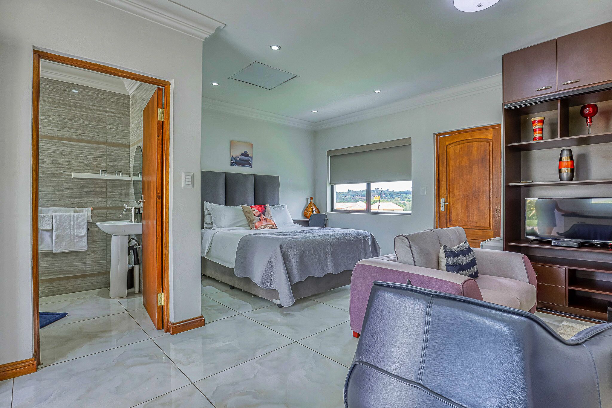 A modern bedroom with a bed, a sofa, a TV, and a bathroom visible through an open door.