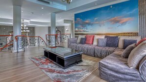 A spacious living room with a large sofa, a glass coffee table, and a mural of a sunset on the wall. - Villa Tasha Luxury suites (Lanseria)