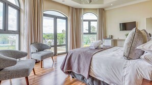 A bedroom with a large bed, two armchairs, a TV, and a balcony with curtains. - Villa Tasha Luxury suites (Lanseria)